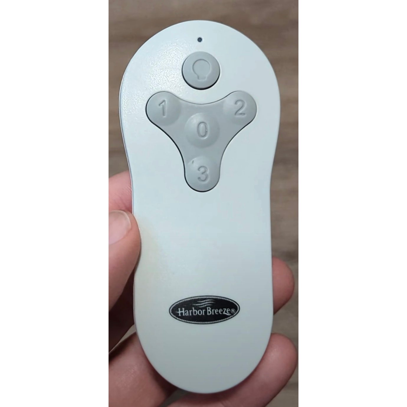 A hand holds a Harbor Breeze TR214B KUJCE10712 ceiling fan remote in white, featuring four gray buttons labeled with a light symbol, 0, 1, and 2. The "Harbor Breeze" logo is at the bottom and FCC ID: TR214B KUJCE10712 is subtly embedded.
