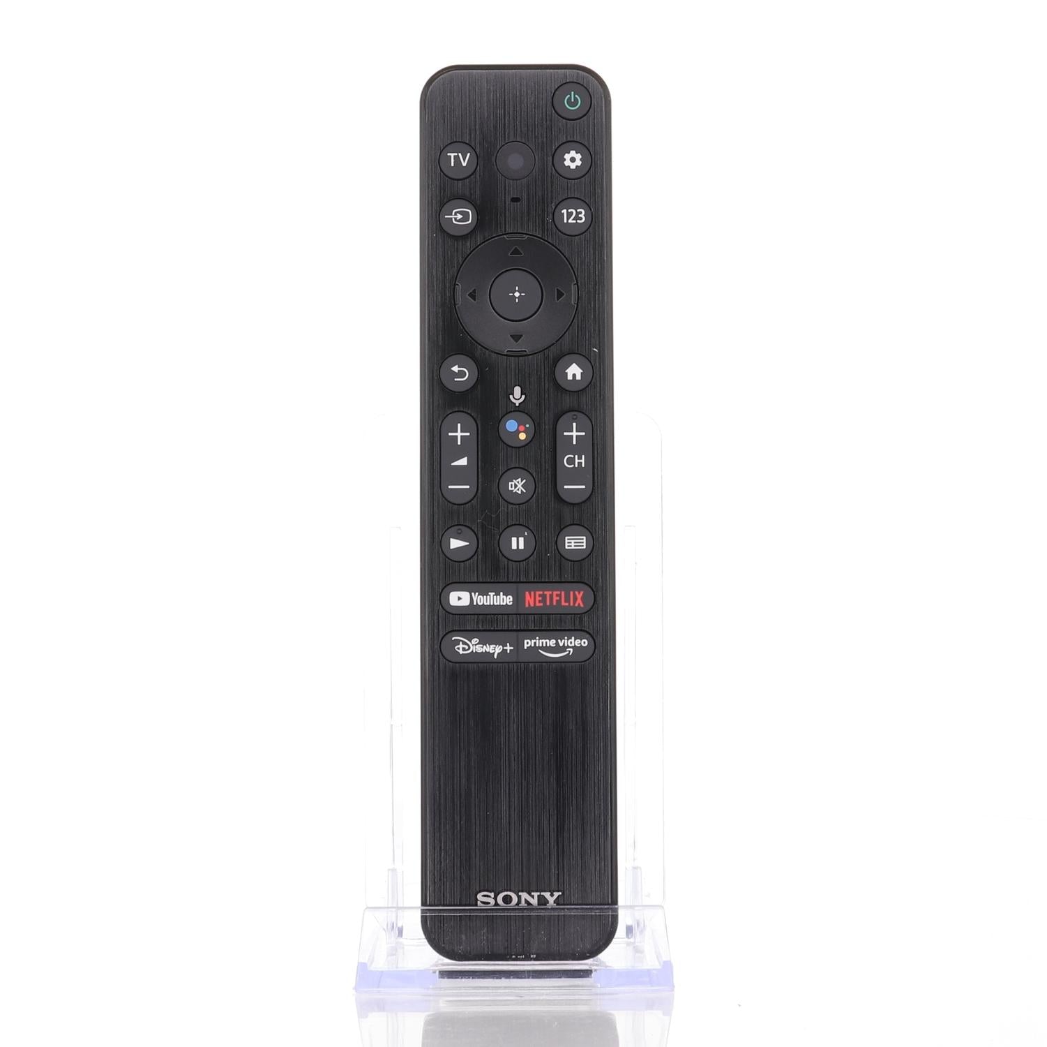The Sony OEM RMF-TX800U RF remote for BRAVIA XR OLED Smart TVs features buttons for YouTube, Netflix, Disney+, and Prime Video, making it a perfect match for your 4K Ultra HDR TV. It is beautifully showcased on a white stand.